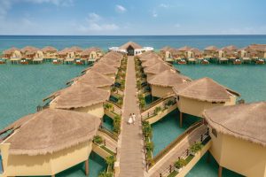 A wide wooden walkway leads to rows of overwater bungalows with thatched roofs built above turquoise ocean water, under a clear blue sky. Two people walk along the central path.