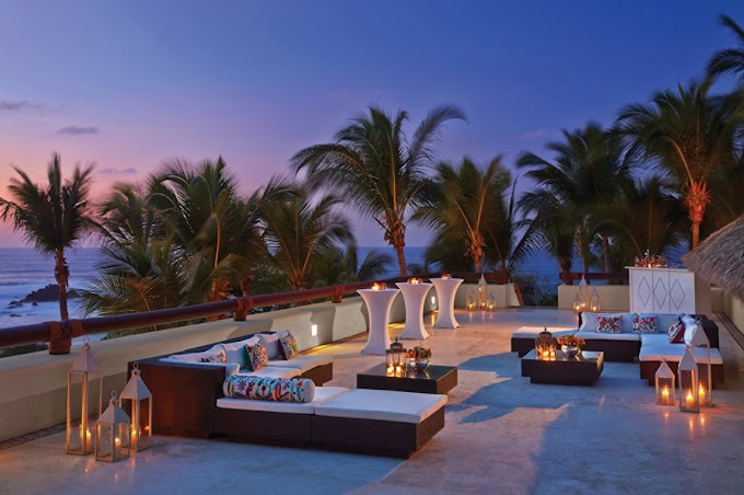 A stylish outdoor lounge with modern seating, lanterns, and candles, set on a terrace overlooking the ocean and palm trees at sunset, creating a warm and inviting atmosphere.