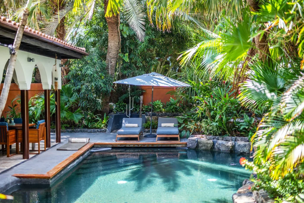 A serene tropical pool area with two lounge chairs under an umbrella, surrounded by lush green plants and palm trees, next to a shaded patio with wooden furniture.