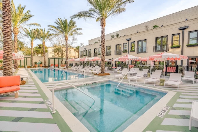 A modern outdoor pool area with palm trees, pink-and-white striped umbrellas, white lounge chairs, and a light-colored building in the background, creating a relaxing resort atmosphere.