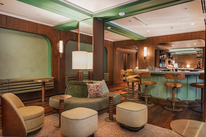 A modern lounge with green and brown decor, cushioned chairs, round tables, a bar with stools, wooden floors, and soft lighting giving the space a cozy, stylish atmosphere.