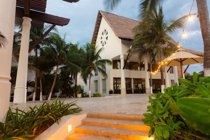 A tropical resort entrance with stone steps, palm trees, string lights, and a white building featuring a thatched roof and large round window, set against a cloudy sky.