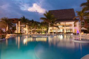A large swimming pool surrounded by palm trees and a thatched-roof resort building is illuminated at dusk, with lights reflecting on the water and a dramatic cloudy sky in the background.