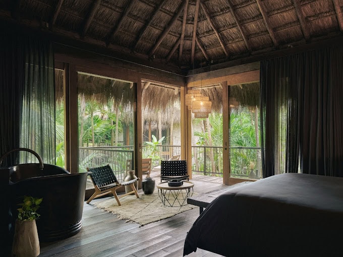 A cozy tropical bedroom with a thatched roof, large windows, and lush greenery outside. The room features a black tub, woven chairs, a small table, and elegant pendant lighting, creating a relaxing and inviting atmosphere.