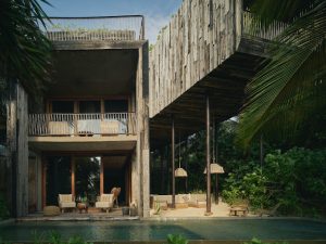 A modern villa with rustic wooden and concrete architecture, large glass doors, a balcony with chairs, an outdoor seating area, and a swimming pool surrounded by lush tropical greenery.