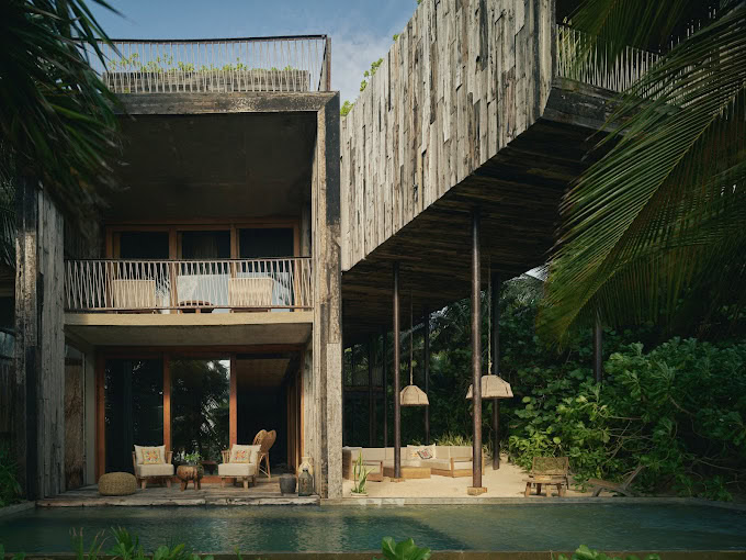 A modern villa with rustic wooden and concrete architecture, large glass doors, a balcony with chairs, an outdoor seating area, and a swimming pool surrounded by lush tropical greenery.