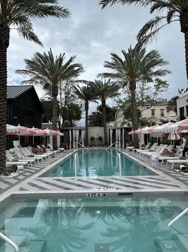 A cloudy day at a stylish outdoor pool lined with palm trees, white lounge chairs, and pink umbrellas. A few people are relaxing by the pool, and buildings are visible in the background.