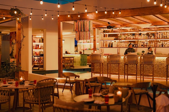 A cozy, warmly lit bar and restaurant with string lights overhead, wicker chairs, round tables set for dining, and a stylish bar area where two people are standing. The atmosphere is inviting and relaxed.