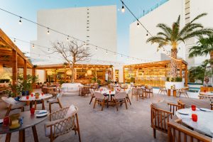 An outdoor restaurant patio with wooden tables and chairs, cozy sofas, string lights overhead, palm trees, and a modern bar area, set against tall white buildings at sunset.