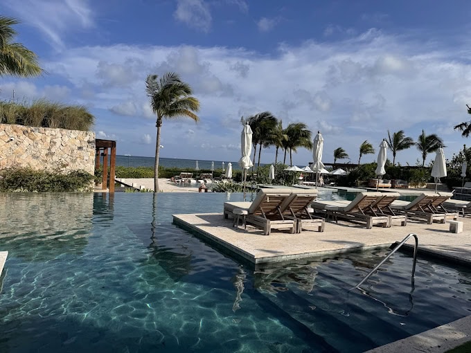 Infinity pool with lounge chairs and umbrellas, surrounded by palm trees, overlooking the ocean under a partly cloudy blue sky. The scene is tropical and relaxing, suggesting a resort or vacation destination.