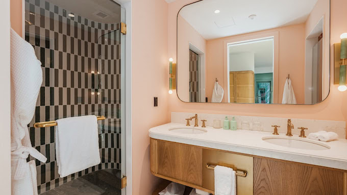 Modern bathroom with a double sink vanity, large mirror, gold fixtures, and a glass shower with black and white tile. White towels and bathrobes are visible, and soft peach walls create a warm atmosphere.
