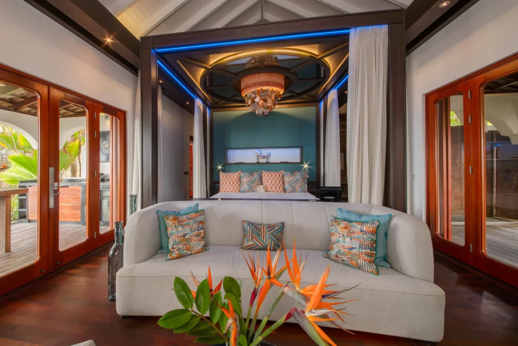 A luxurious, modern bedroom features a plush light-gray sofa with colorful pillows, a canopy bed with accent lighting, wooden floors, and large glass doors opening to a tropical outdoor view. Bird of paradise flowers are on a table in front.