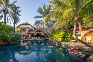 A luxurious villa with terracotta roofs and arched doorways, surrounded by palm trees and lush greenery, overlooks a clear blue swimming pool with lounge chairs and a small toy boat floating on the water.