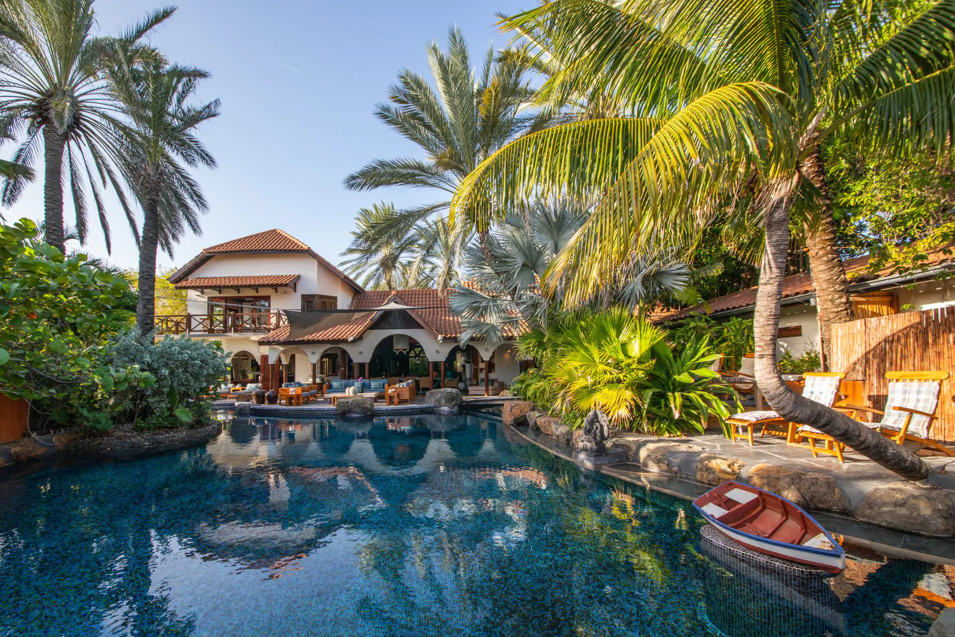 A luxurious villa with terracotta roofs and arched doorways, surrounded by palm trees and lush greenery, overlooks a clear blue swimming pool with lounge chairs and a small toy boat floating on the water.