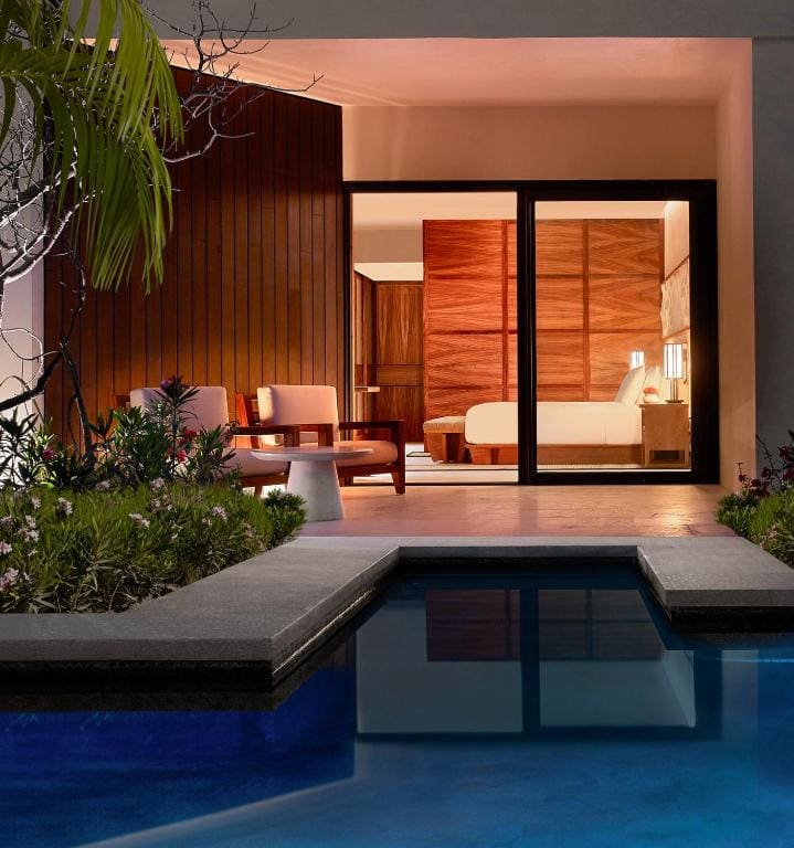 A view of a modern hotel room with large glass doors opening to a private patio, outdoor seating, lush plants, and a blue swimming pool in the foreground. Warm lights illuminate the cozy interior and exterior spaces.