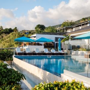 A modern outdoor pool with clear blue water, surrounded by lounge chairs and blue umbrellas, set against a backdrop of greenery and a hillside under a partly cloudy sky.