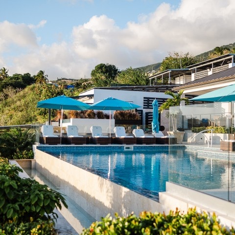 A modern outdoor pool with clear blue water, surrounded by lounge chairs and blue umbrellas, set against a backdrop of greenery and a hillside under a partly cloudy sky.