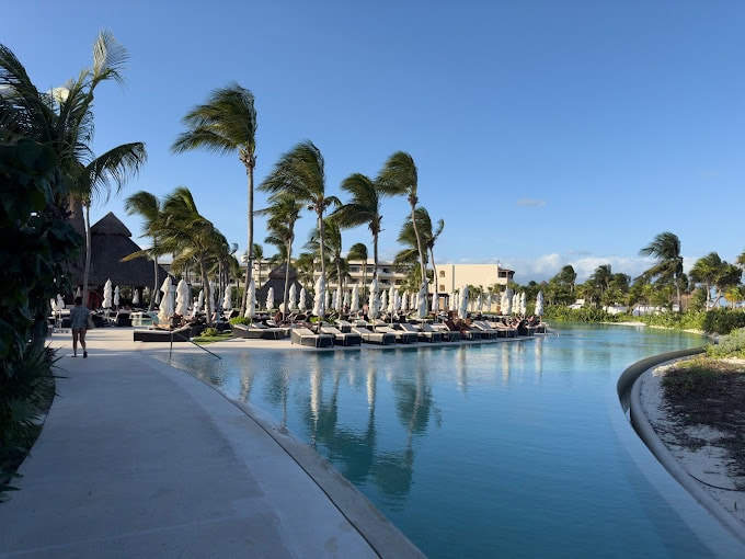 A resort swimming pool is lined with lounge chairs and umbrellas, bordered by tall palm trees swaying in the wind under a clear blue sky. A few people relax poolside, enjoying the sunny tropical atmosphere.
