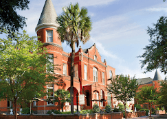 Historic red brick building with a pointed turret, arched windows, and lush green trees in the foreground under a blue sky.