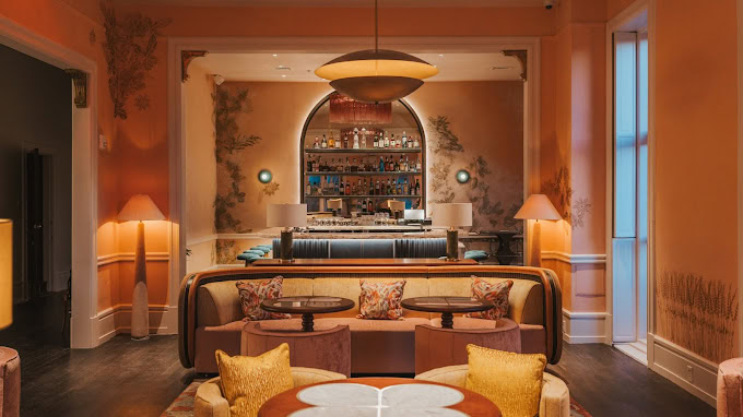 Stylish lounge with peach-colored walls, cozy velvet seating, round tables, soft lighting, and a bar in the background stocked with bottles and glassware. Elegant lamps and artistic wall décor create a warm, inviting atmosphere.