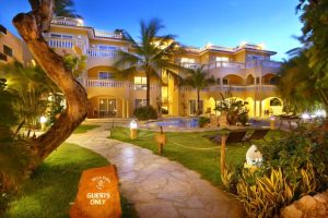 A well-lit resort building at dusk features palm trees, a winding stone path, a pool, lounge chairs, and a sign reading Guests Only. Lush greenery surrounds the tranquil, inviting setting.