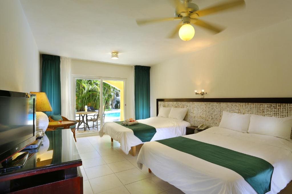 A bright hotel room with two double beds, white bedding and green runners, a TV, a desk, a lamp, ceiling fan, and sliding glass doors opening to a patio with chairs and a view of a pool and palm trees.