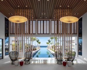 Modern indoor lounge with wicker chairs and pendant lights, featuring wooden slats and large windows overlooking a pool, palm trees, and the ocean in the background.