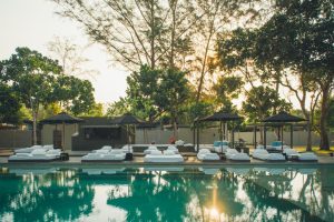 A serene outdoor pool area with white lounge beds and black umbrellas, surrounded by trees and greenery, with sunlight filtering through the branches in the background.