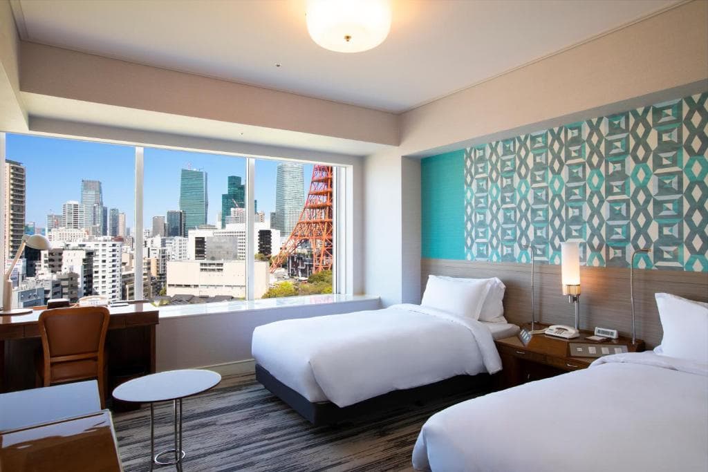 A modern hotel room with two white beds, a desk and chair by a large window, and patterned blue wallpaper. The window offers a city view with buildings and a prominent red tower outside.