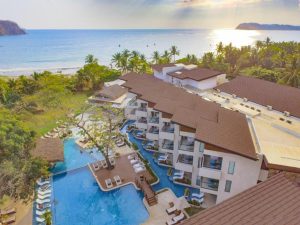 Aerial view of a modern beachfront resort with a large pool, sun loungers, palm trees, and direct access to the sandy beach and ocean under a bright, sunny sky.