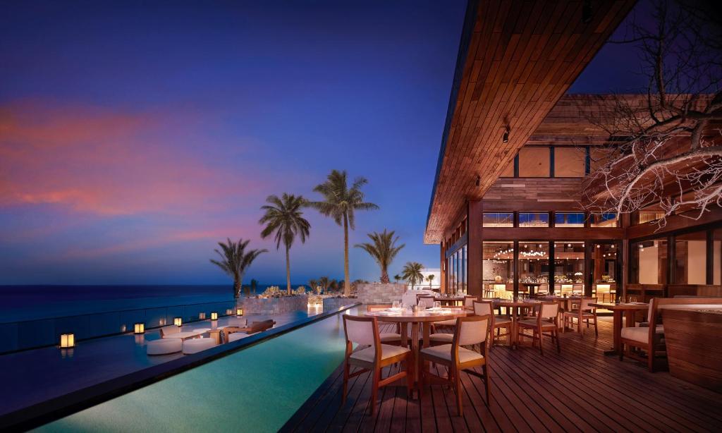 A luxurious outdoor restaurant with wooden tables and chairs overlooks an infinity pool, palm trees, and the ocean at sunset, with soft lighting creating a serene and inviting atmosphere.