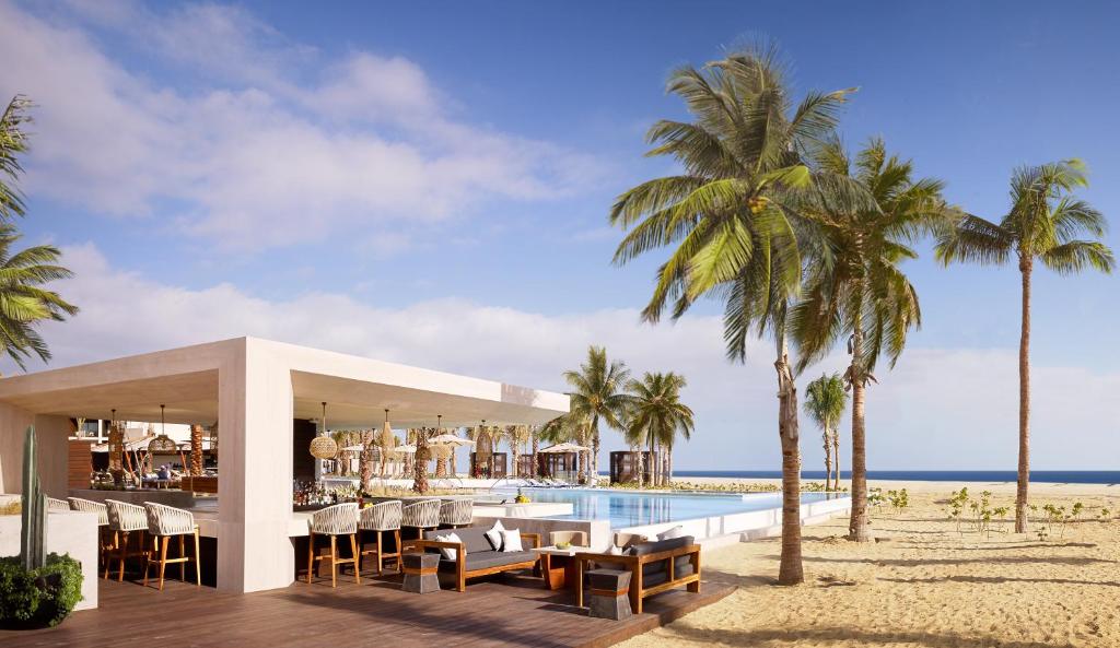 Modern beachside bar and lounge with wooden furniture and hanging lights, next to a pool, surrounded by palm trees on sandy ground, with the ocean and blue sky in the background.