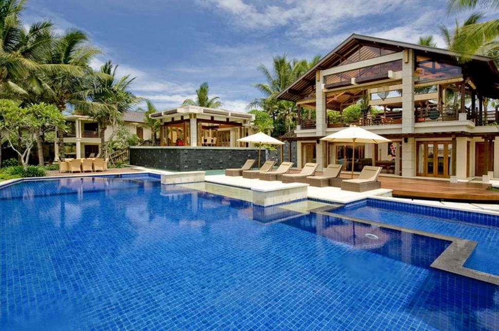Luxury villa with wooden accents, large blue-tiled swimming pool, sun loungers, poolside umbrellas, and tropical palm trees, under a bright blue sky.