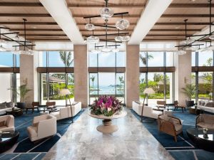 A spacious, modern hotel lobby with high ceilings, large windows, elegant seating areas, and a round centerpiece table displaying vibrant purple flowers. Tropical scenery and ocean views are visible through the windows.