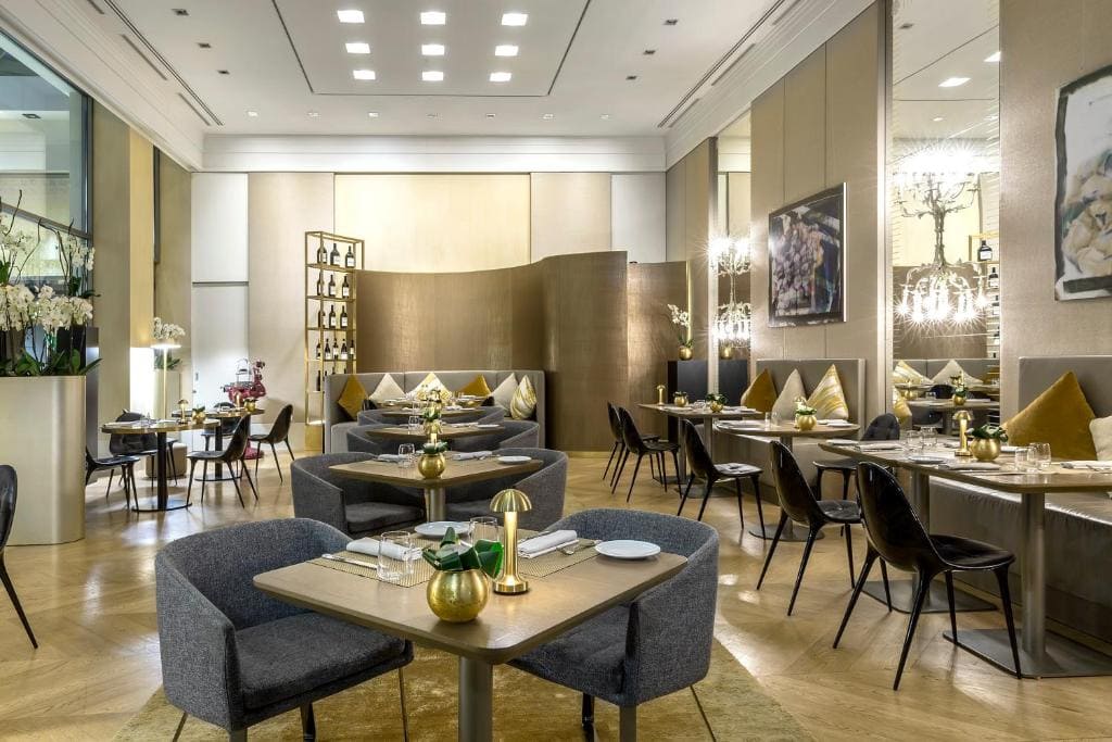 Modern, elegant restaurant interior with gray and gold decor, cushioned chairs, stylish lighting, and set tables. Partition walls, artwork, and potted plants create a sophisticated, inviting dining atmosphere.