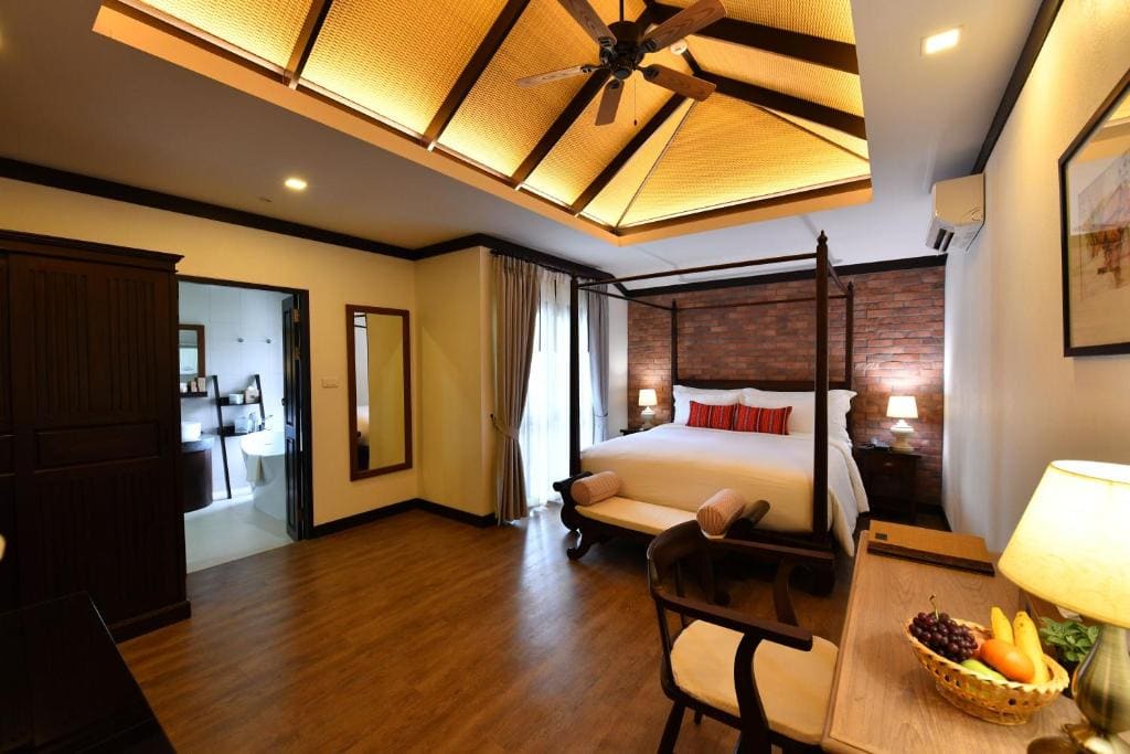 A cozy hotel room with a wooden floor, canopy bed, brick accent wall, and warm lighting. There’s a desk with a chair and fruit bowl, a wardrobe, and an open bathroom visible through a door. Ceiling fan and lamps complete the decor.