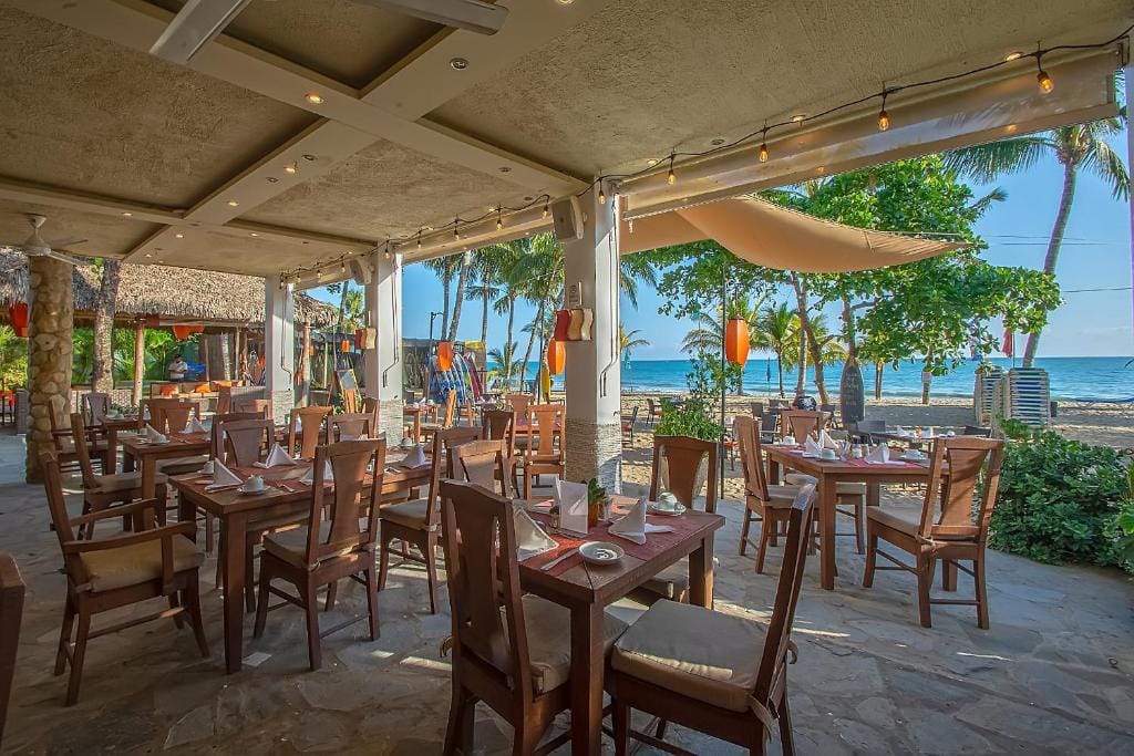 Open-air seaside restaurant with wooden tables and chairs set for dining, tropical trees and plants surrounding the area, and a clear view of the sandy beach and blue ocean in the background.