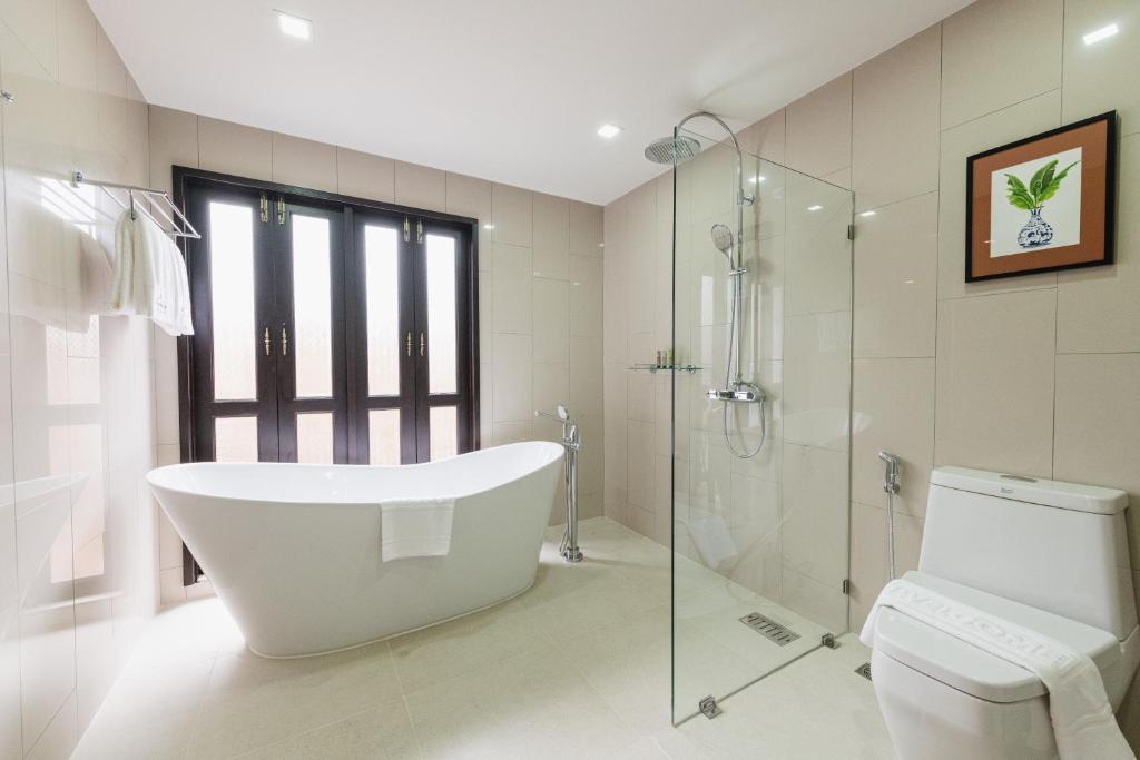 Modern bathroom with beige tiles, a freestanding white bathtub by large glass doors, a glass-enclosed shower, a toilet, towel rack, and a framed plant picture on the wall. The space is bright and minimalistic.