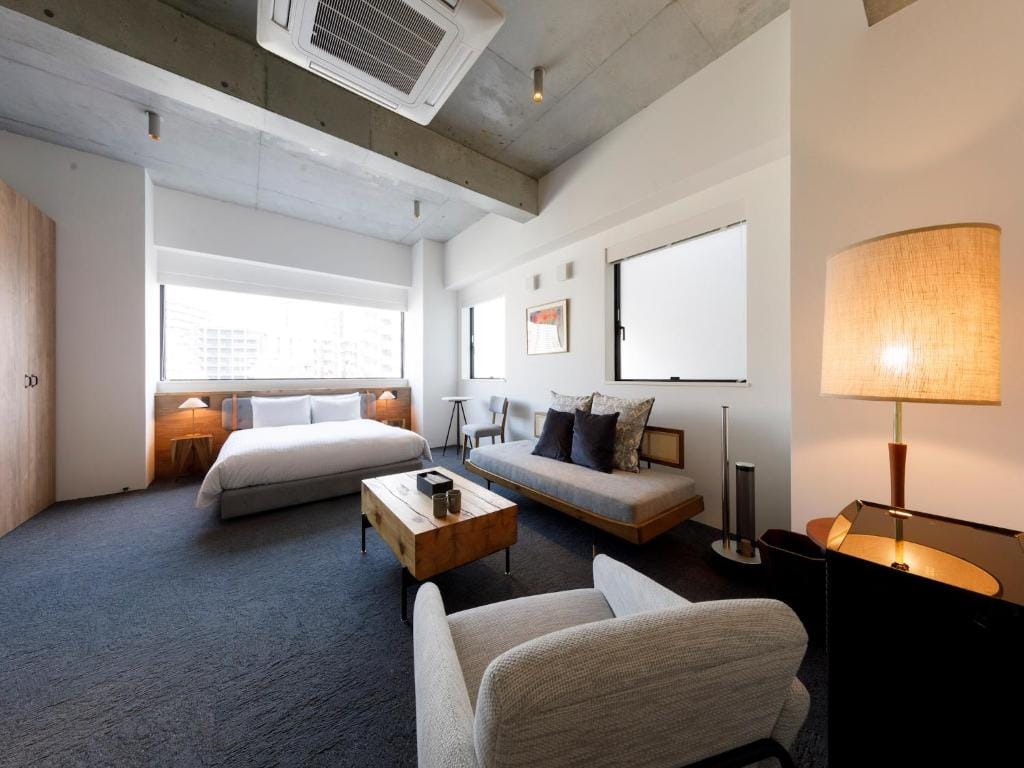 A modern, minimalist bedroom with a large bed, gray sofa, armchair, wooden coffee table, two nightstands, lamps, and large windows letting in natural light. The ceiling is exposed concrete with a visible air conditioning unit.