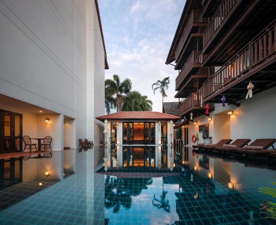 A serene hotel courtyard with a turquoise tiled swimming pool, lounge chairs, and wooden balconies. Palm trees and a poolside pavilion are visible, with reflections on the still water under a partly cloudy sky.