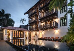 Modern hotel with balconies overlooking a pool lined with lounge chairs, surrounded by palm trees, illuminated in the evening. A separate building with large windows is visible on the left.