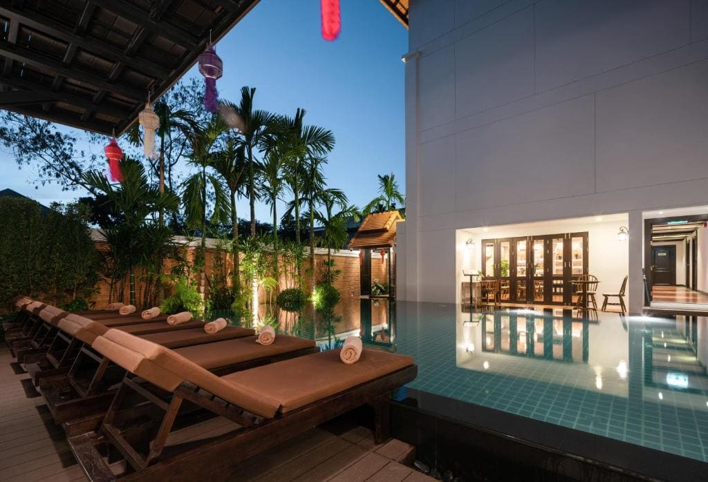 Outdoor pool area at dusk with wooden lounge chairs and rolled towels, palm trees, lanterns, and a modern building with large windows and warm lights reflecting in the water.