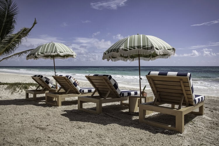 Four wooden lounge chairs with blue-and-white striped cushions sit on a sandy beach under green-striped umbrellas, facing the ocean. A palm tree is visible to the left, and the sky is partly cloudy.