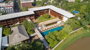 Aerial view of a modern hotel complex with an L-shaped building, central courtyard, long swimming pool, surrounding greenery, and a river alongside the property. Adjacent city buildings and trees are visible.