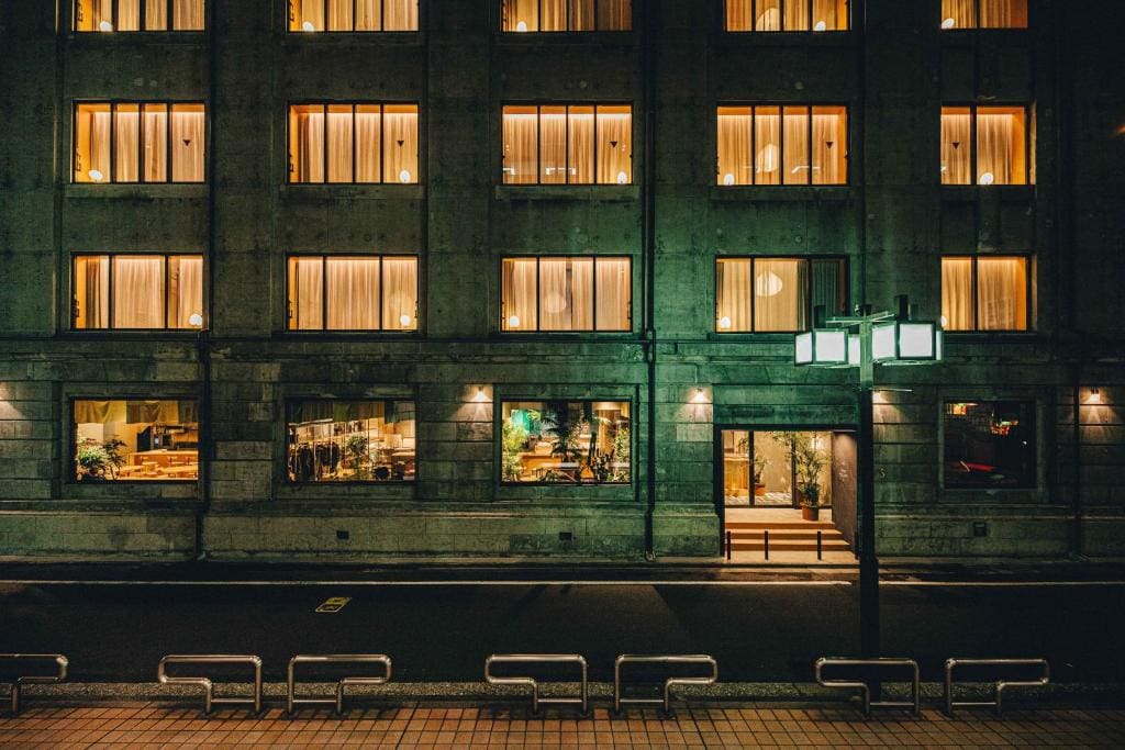 A warmly lit building at night features large windows with glowing lights and curtains. The ground floor reveals a cozy restaurant interior. Empty benches and a streetlamp line the quiet street outside.
