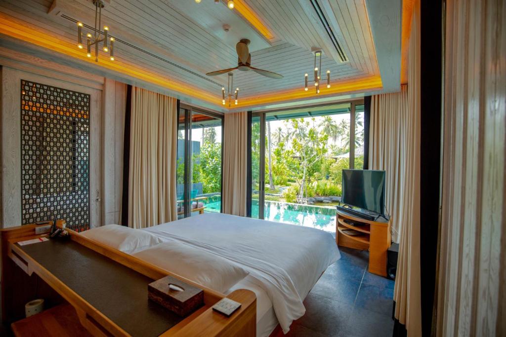 A modern hotel room with a large bed, wooden furnishings, ceiling fan, and floor-to-ceiling windows that open to a private pool and lush garden view, allowing natural light to fill the space.