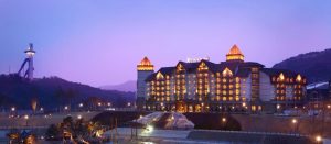 A large, grand hotel with illuminated windows and roof peaks stands against a dusky purple sky, surrounded by mountains; a ski jump tower is visible to the left.