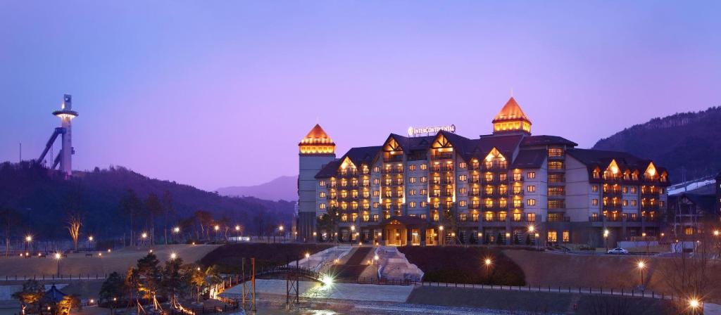 A large, grand hotel with illuminated windows and roof peaks stands against a dusky purple sky, surrounded by mountains; a ski jump tower is visible to the left.