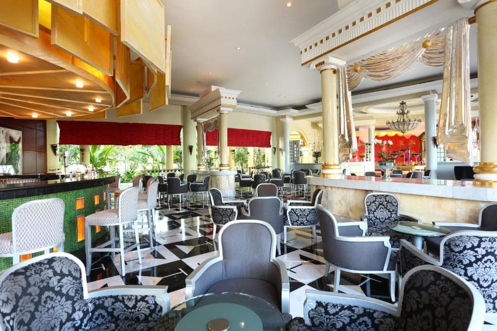 Elegant hotel lounge with black and white patterned chairs, round glass tables, marble columns, checkered floor, and large windows with red curtains, featuring ornate gold decor and greenery outside.