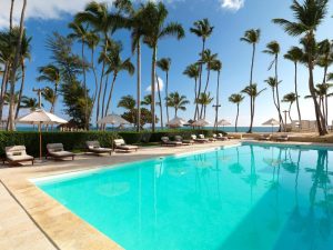 A serene outdoor pool surrounded by lounge chairs and umbrellas, with tall palm trees and a clear blue sky in the background, overlooking a sandy beach and the ocean.